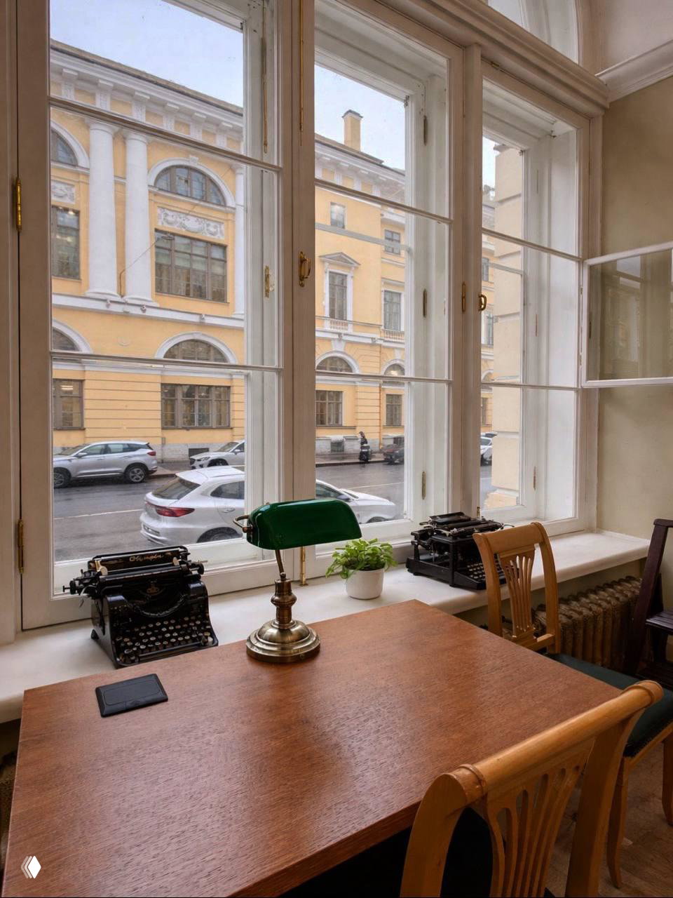 Рабочее место у большого окна библиотеки: деревянный стол с зелёной лампой и двумя старинными печатными машинками, вид на фасад по ул. Зодчего Росси.