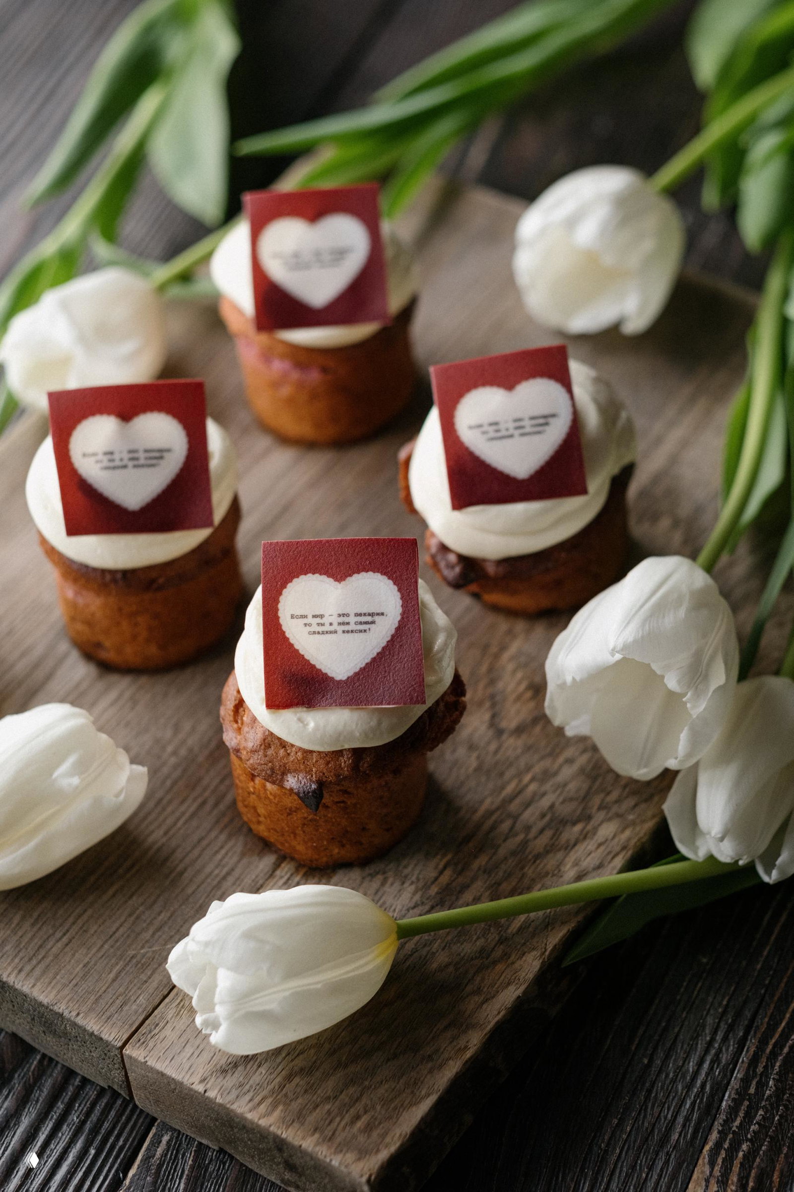 Набор небольших малиновых кексов с белым кремом и съедобными сердечками, выложенные на деревянной доске рядом с белыми тюльпанами — спешл-десерт для праздника.