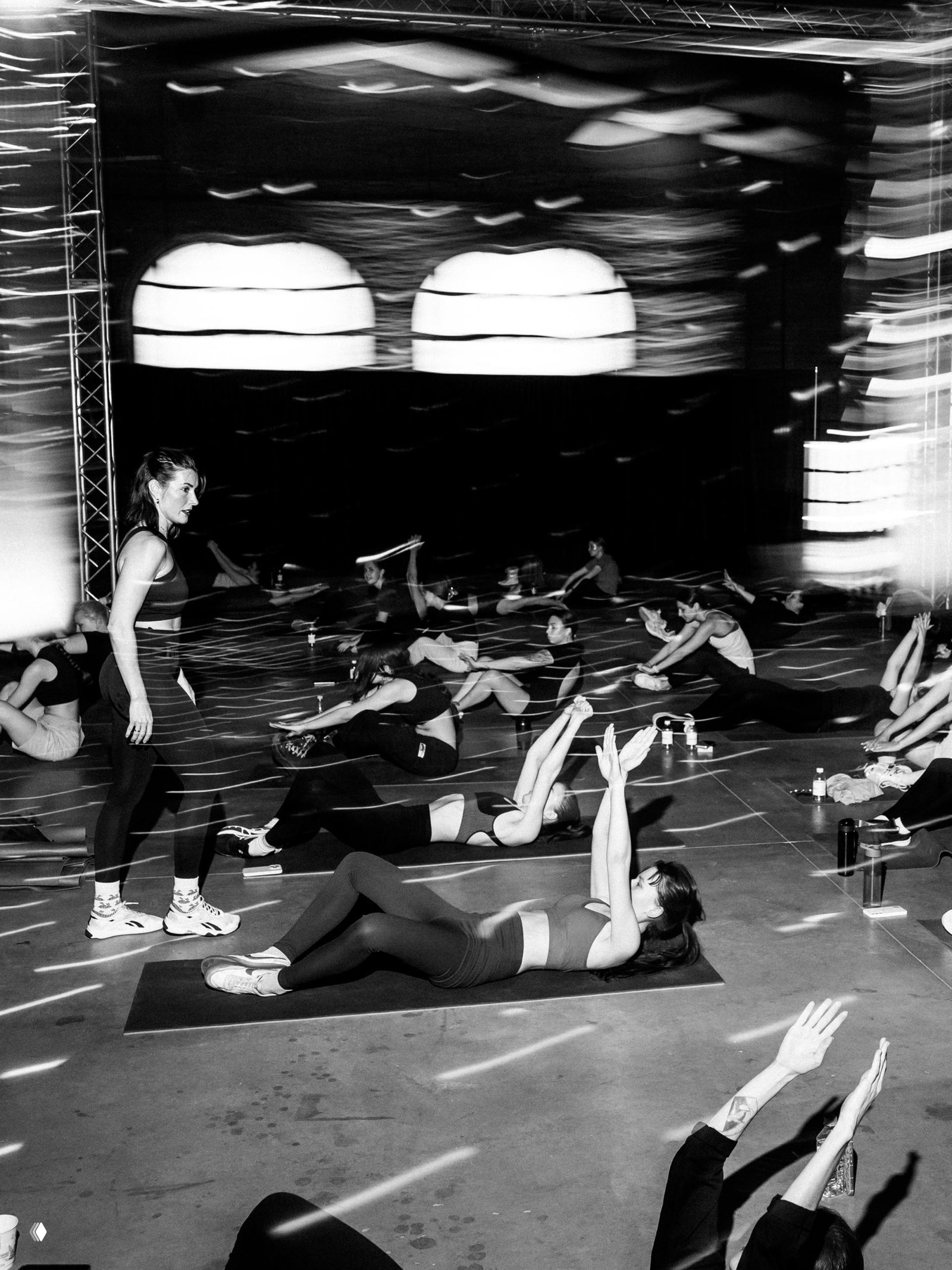 Черно‑белая фотография групповой тренировки: участники на ковриках выполняют упражнения в большом зале под эффекты света.