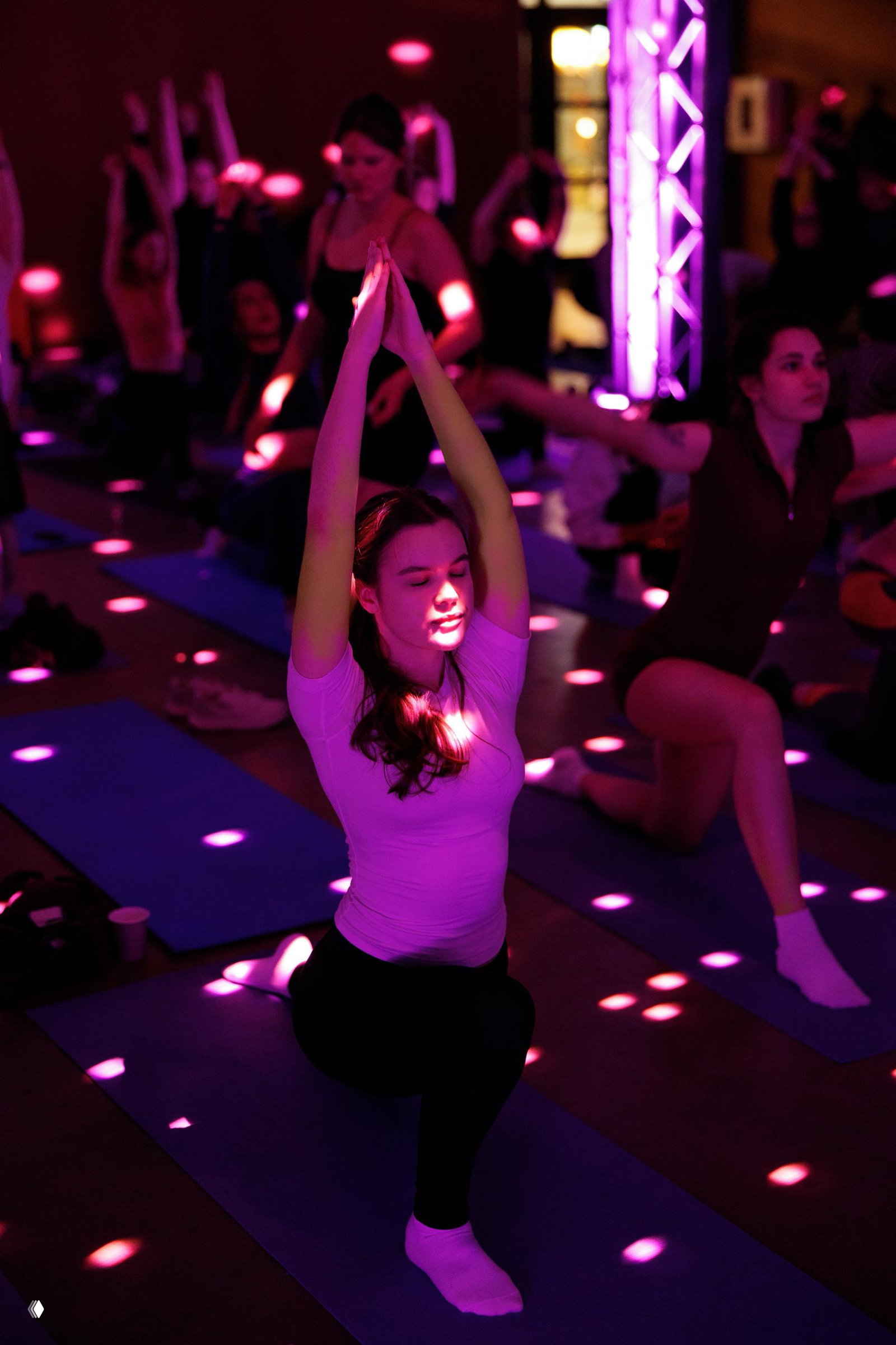Цветная фотография участницы в позе йоги на коврике при пурпурной подсветке и точечных световых эффектах в зале.