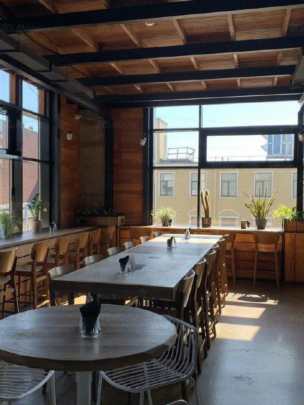 Просторный зал с большими окнами и длинными столами, много естественного света, идеальное место для групповой работы или удалённого офиса на день.