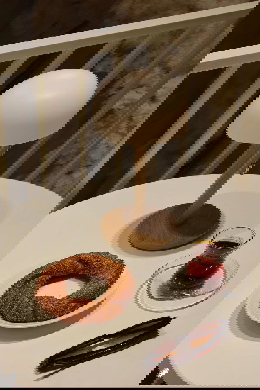 На столе — свежий симит и стакан турецкого чая рядом с солнцезащитными очками и миниатюрной лампой; типичная подача выпечки и напитков в Nerede.