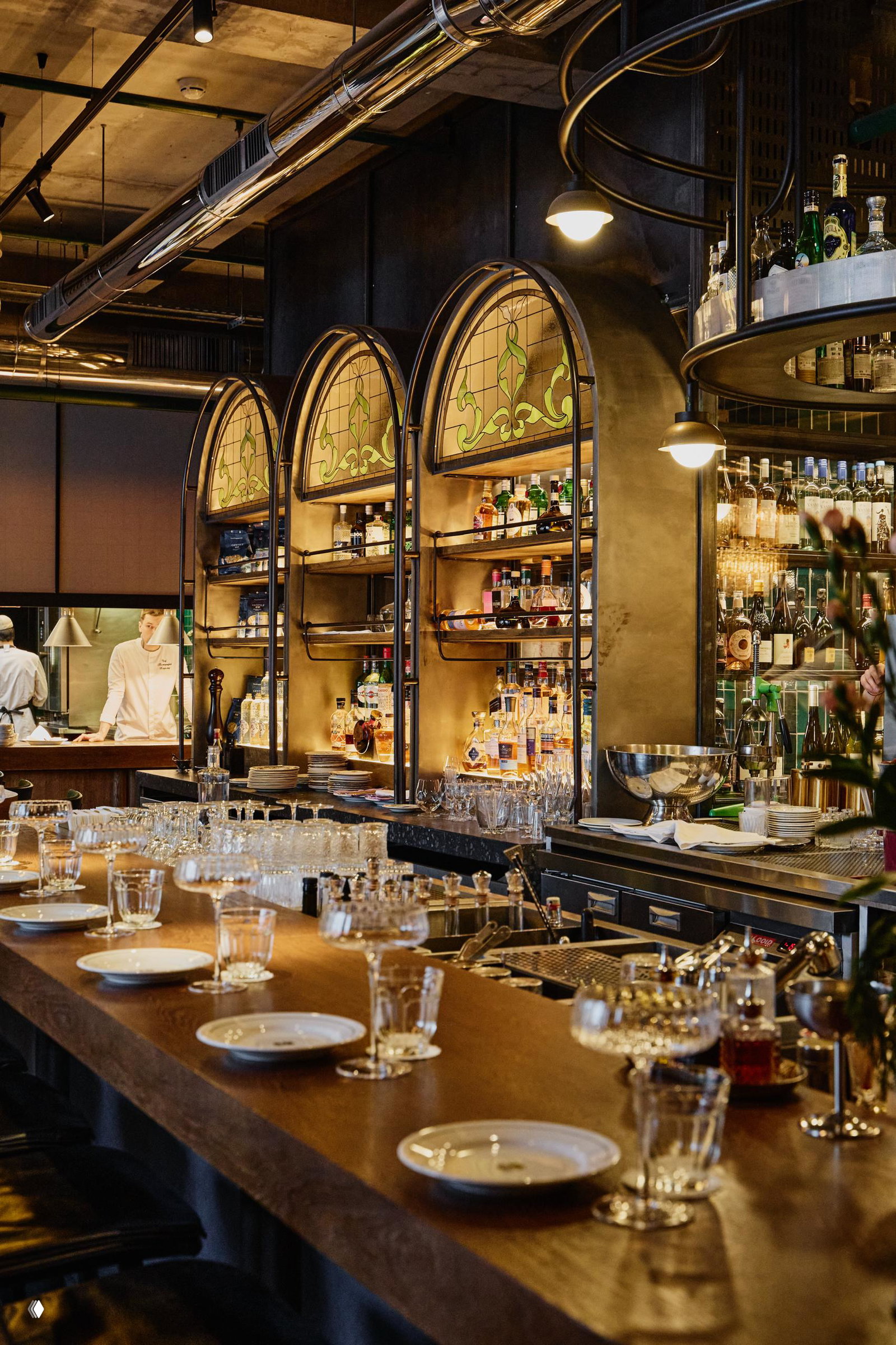 Интерьер бара Bist: длинная стойка сервирована бокалами и тарелками, полки с бутылками и теплое освещение — атмосфера перед вечеринкой.