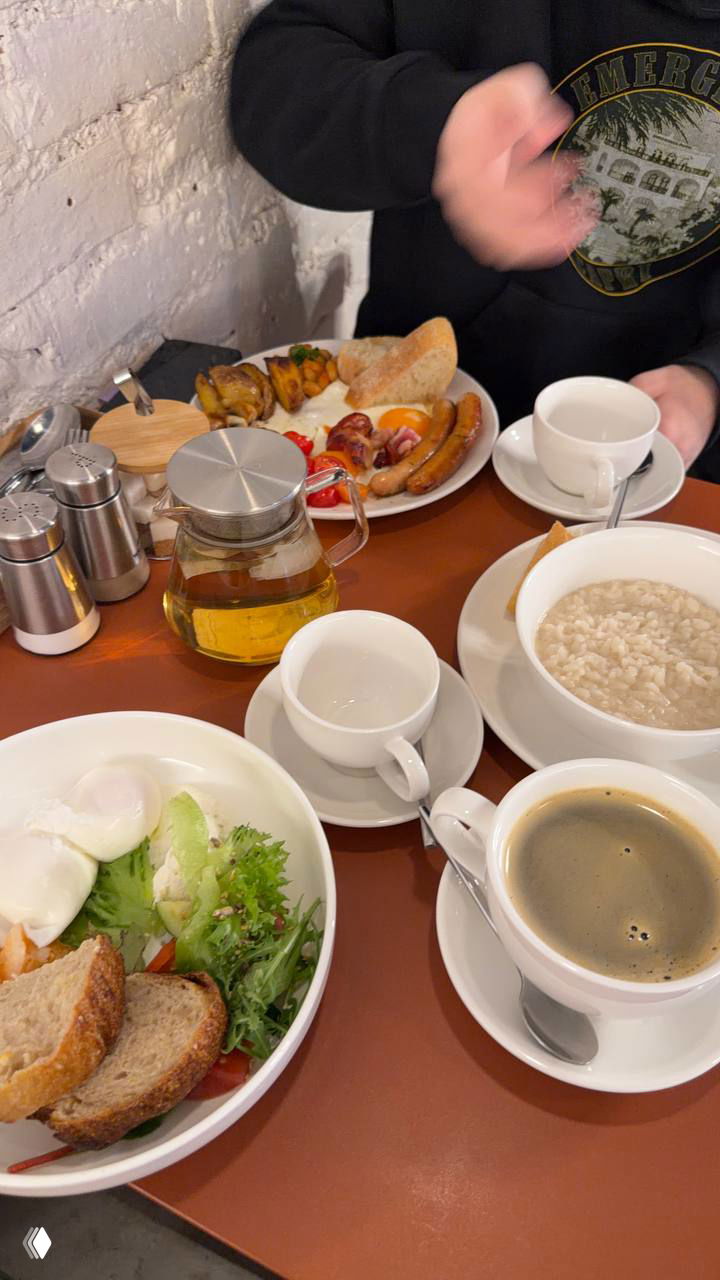 Вид сверху на стол с несколькими завтраками: овсянка, сосиски, хлеб, салат, чашки кофе и чайник — общий снимок утренней подачи в кафе.