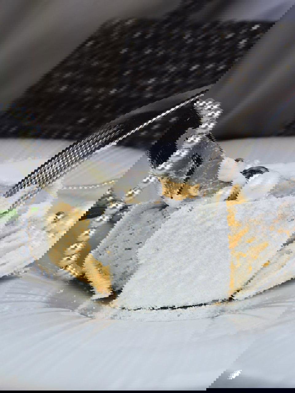 Кусочек кокосового десерта с жемчужными украшениями, разрезанный вилкой на фоне текстурной сумки и бус.