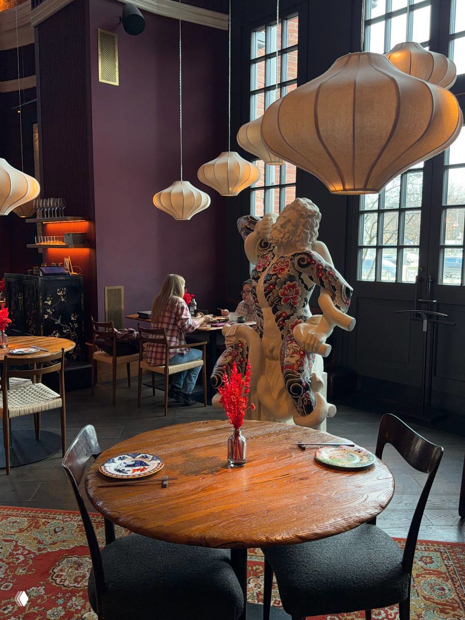 Общий вид зала ресторана: круглые столы, большие текстильные подвесные лампы и декоративная скульптура у окна — светлая крупная витрина и уютная гостиная атмосфера.