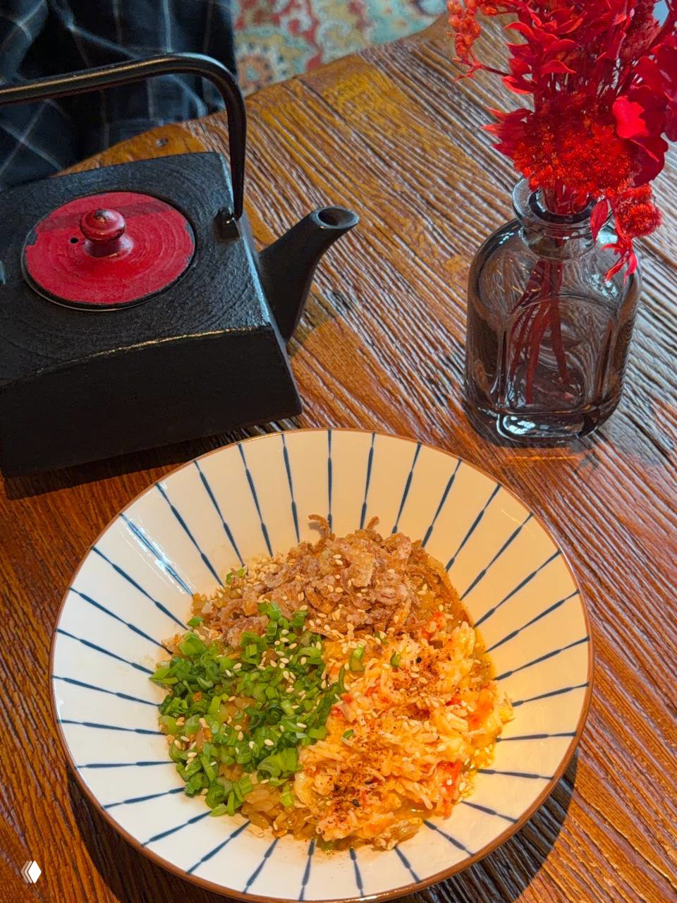 Тарелка с жареным рисом с крабом, украшенным зелёным луком и хрустящими хлопьями, рядом тёмный чайник и красные сухие цветы — аппетитная подача блюда.