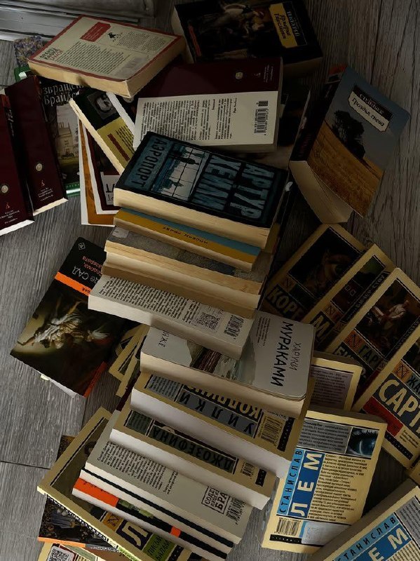 Стопки и разбросанные книги на деревянном полу: видны обложки и корешки разных изданий, среди них книги на русском и имя Мураками на одной из обложек.