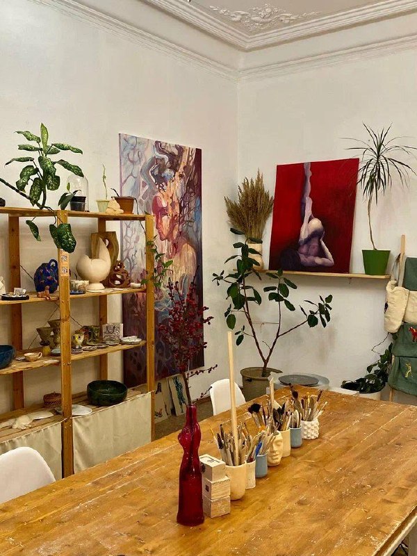 Творческая студия: деревянный стол с кистями, полки с керамикой и картины на стене — интерьер мастерской для лепки.