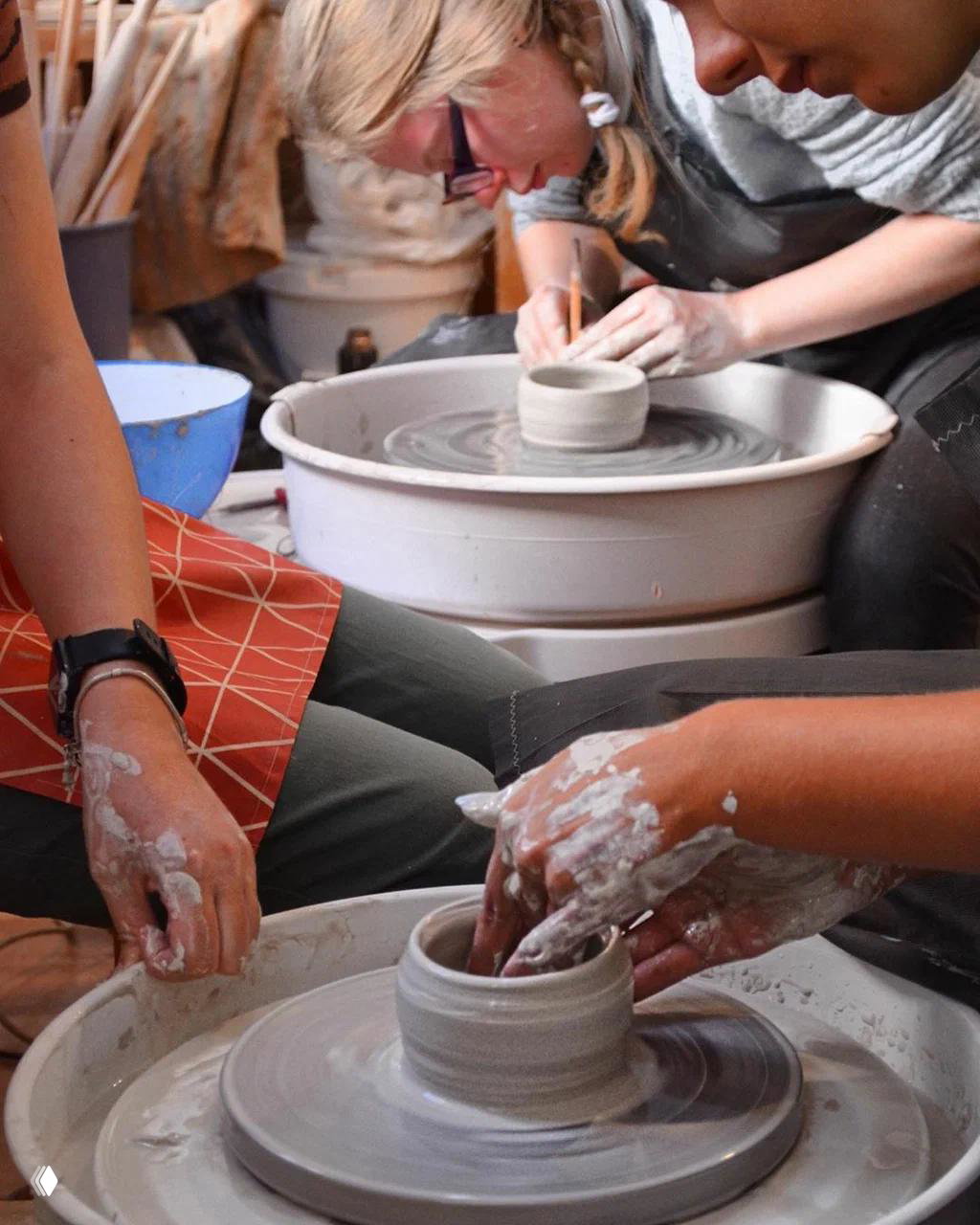 Близкий план: участники за гончарным кругом формуют изделие из глины, руки покрыты глиной — демонстрация процесса мастеркласса.