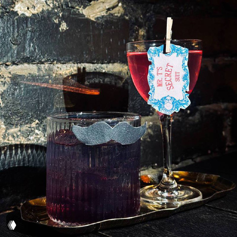 Два коктейля на подносе у кирпичной стены: высокий бокал с биркой «Mr. T's Secret» и низкий стакан с усиком‑декором, красивая подача напитков.