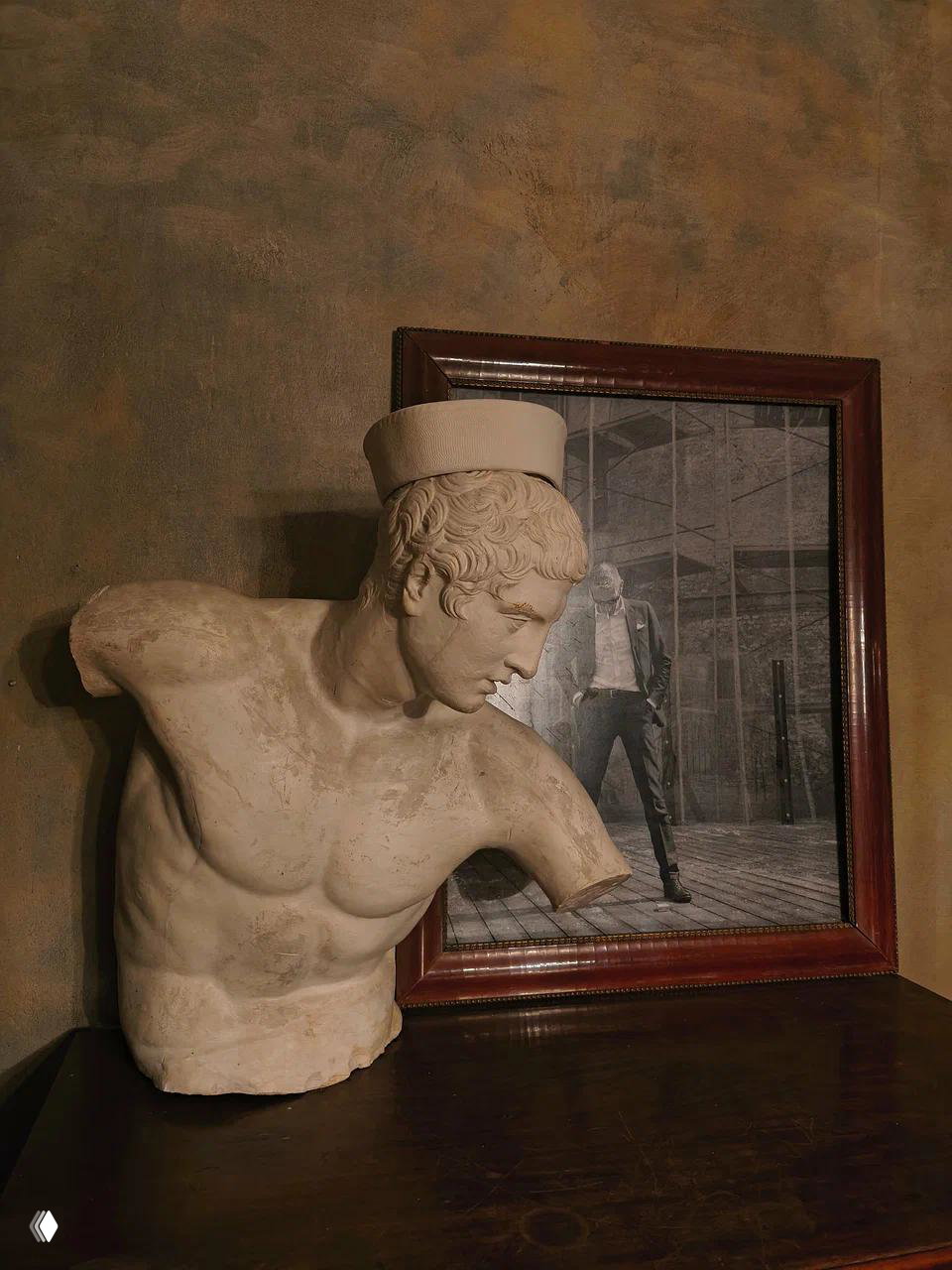 Экспонаты в музее: мраморный торс‑бюст на тумбе на фоне рамной чёрно‑белой фотографии и текстурной стены интерьера.