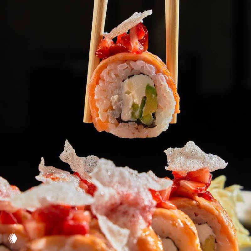 Крупный план ролла, удерживаемого палочками: аккуратный рис, начинка с авокадо и крем‑чизом, сверху хрустящие декорации и кусочки красных ягод.