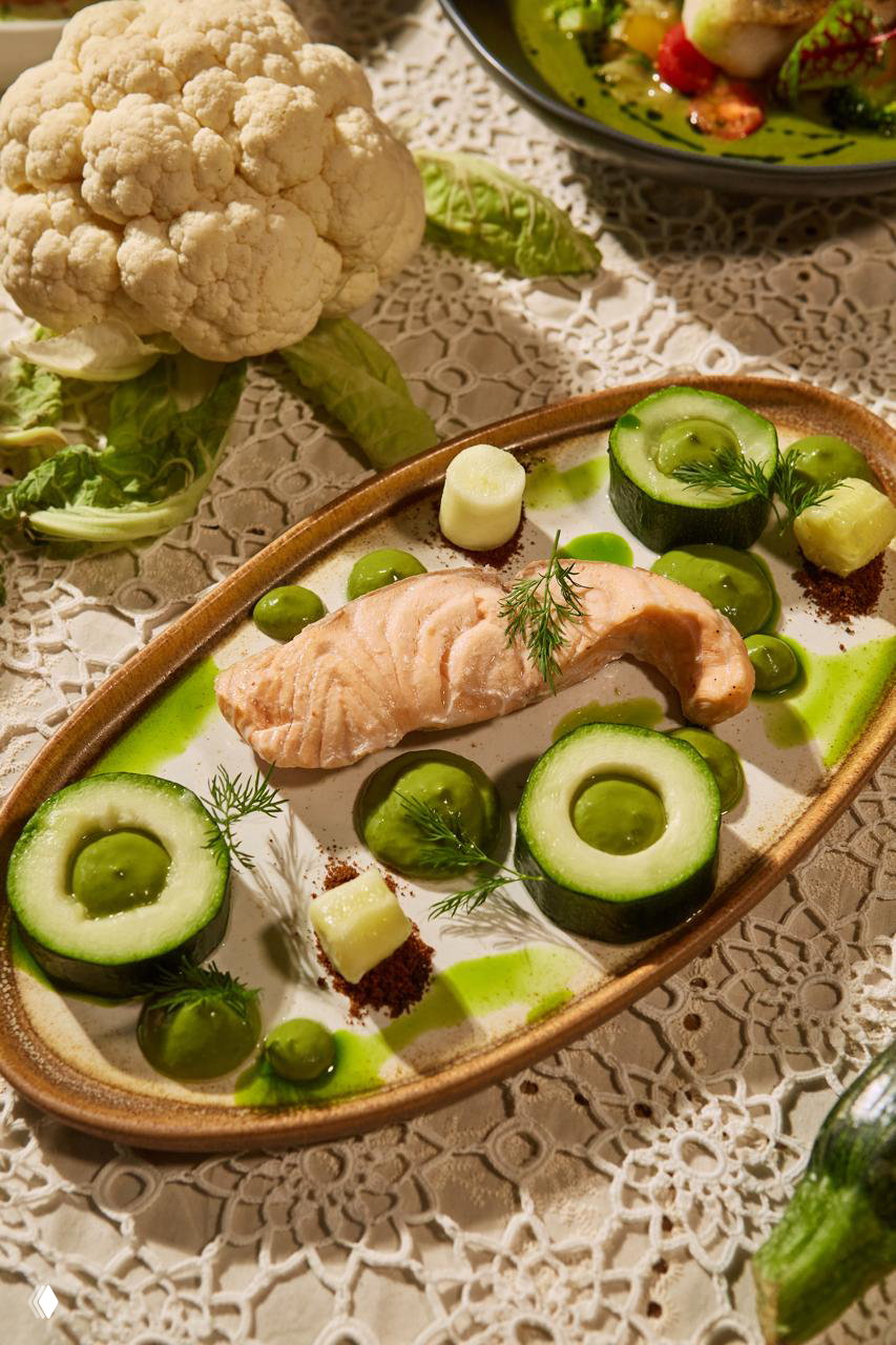 Тарелка со слабосолёной неркой и декоративными кругами огурца, зелёными соусами и кусочками цветной капусты на текстильной салфетке в летней сервировке.