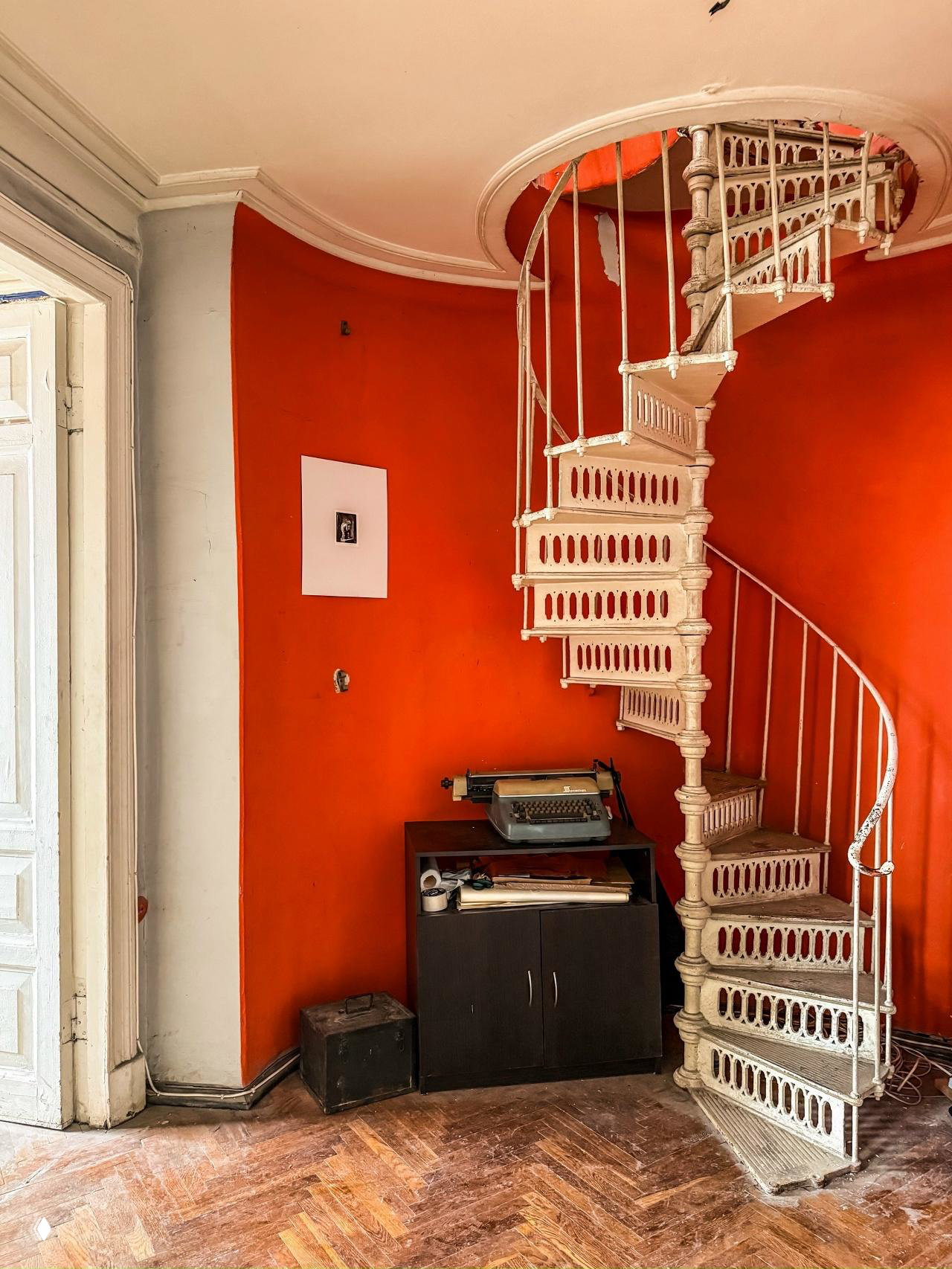 Кованая винтовая лестница в интерьере мастерской на фоне насыщенно‑оранжевой стены, рядом старый пишущий аппарат и предметы быта на тумбе.