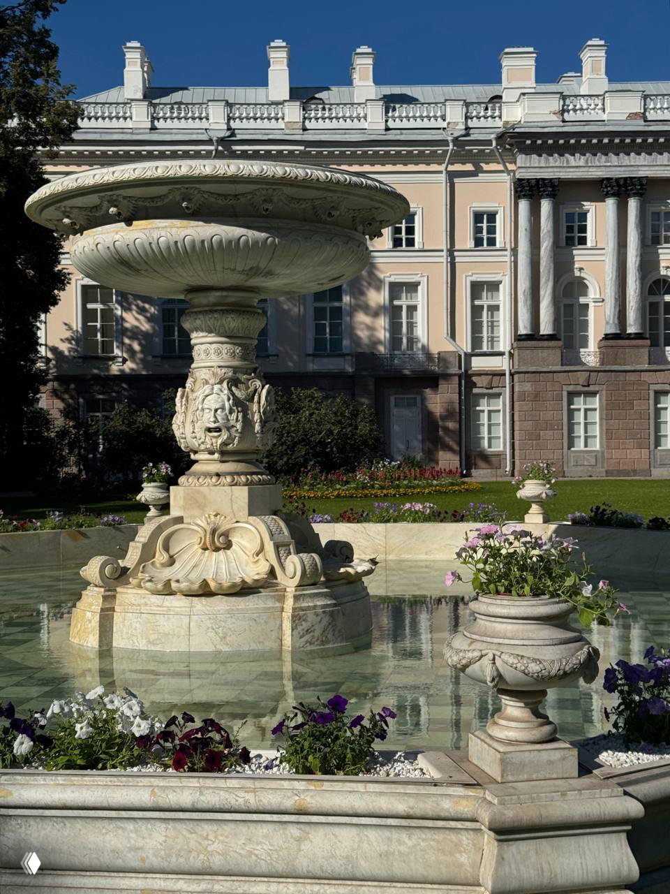 Мраморный фонтан в парке на фоне фасада дворца: вода, цветочные клумбы и игра света создают камерную летнюю атмосферу.