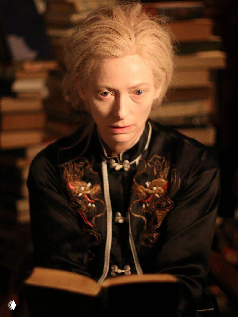 Крупный план актрисы с бледной кожей, читающей книгу; тёплый свет и фоновая стена со стопками книг создают интимную сцену.