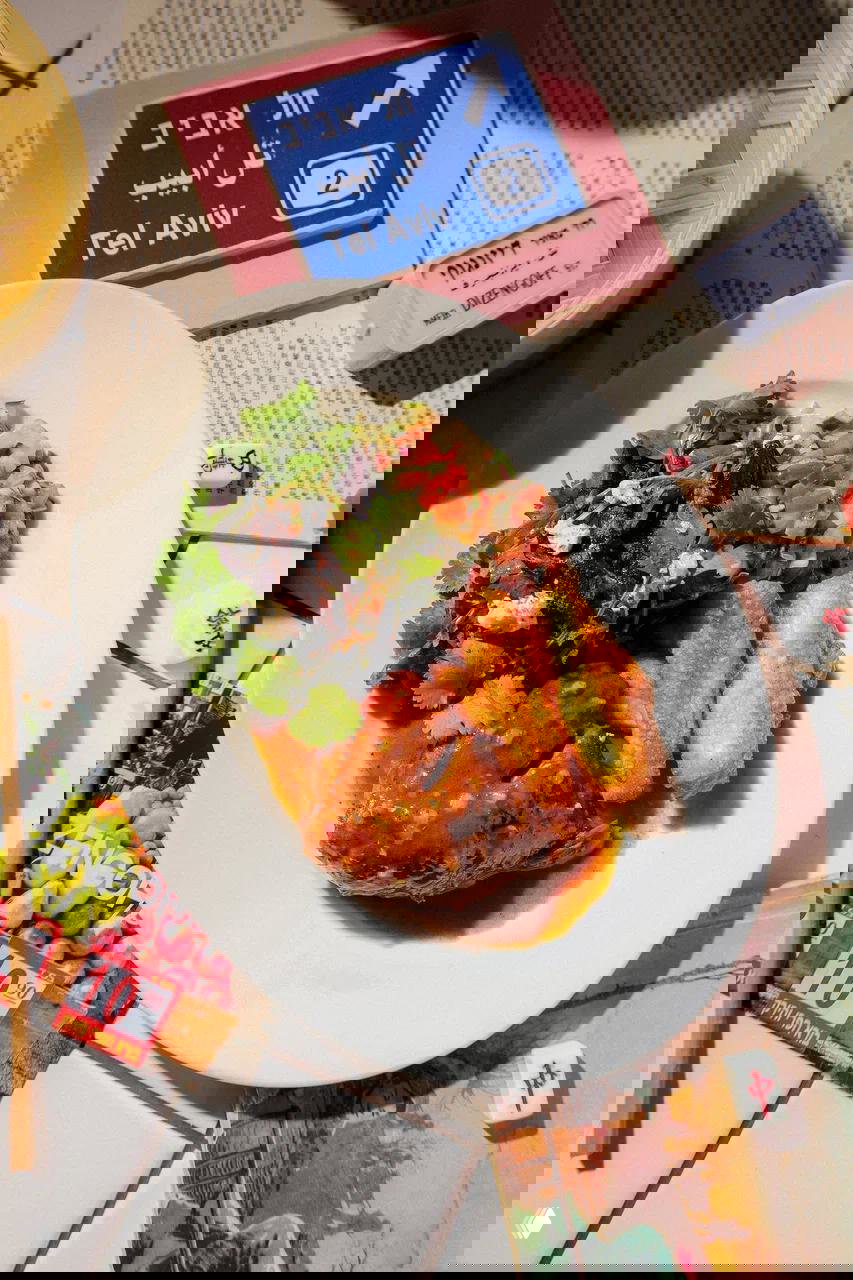 Крупный план блюда: томлёная баранина с жареным халуми, зелёный салат и соус на фоне столешницы с табличкой Tel Aviv и газетами.