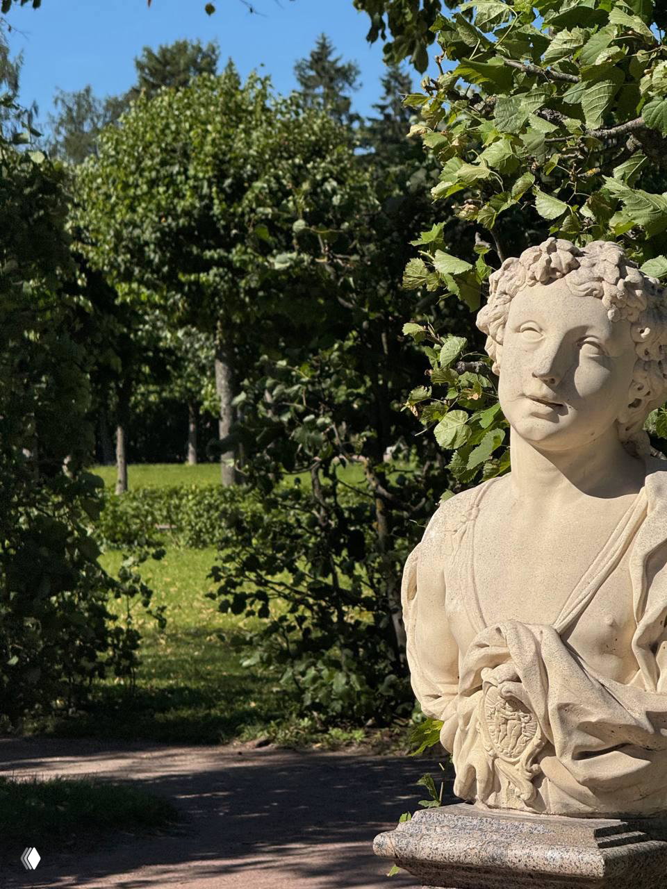 Каменная классическая скульптура в парковой аллее на фоне густой зелени: бюст в солнечном боковом свете, аккуратная садовая композиция.