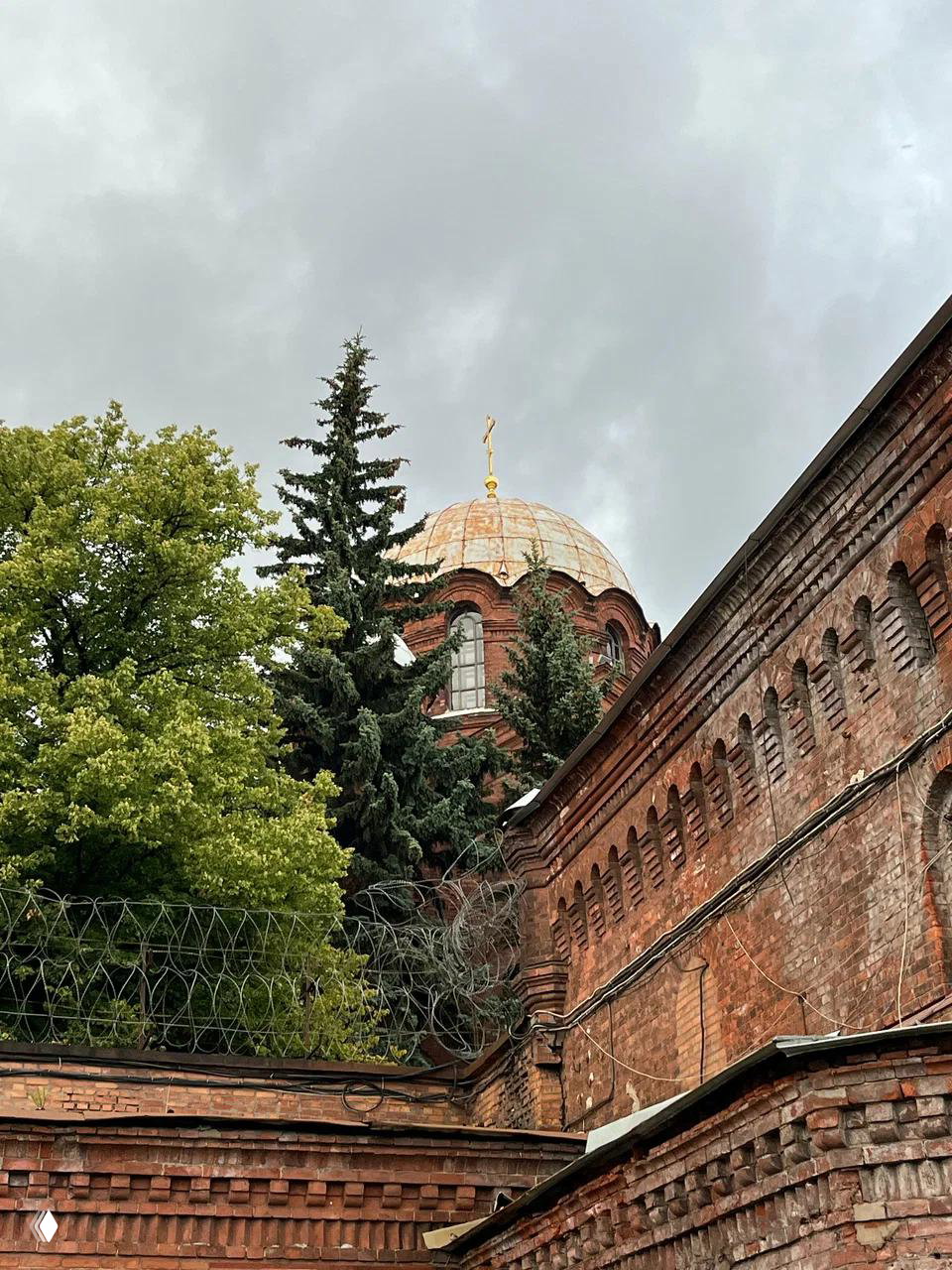Внешний вид кирпичного корпуса «Кресты»: купол часовни виднеется за стеной, колючая проволока и зелёные деревья на фоне пасмурного неба.