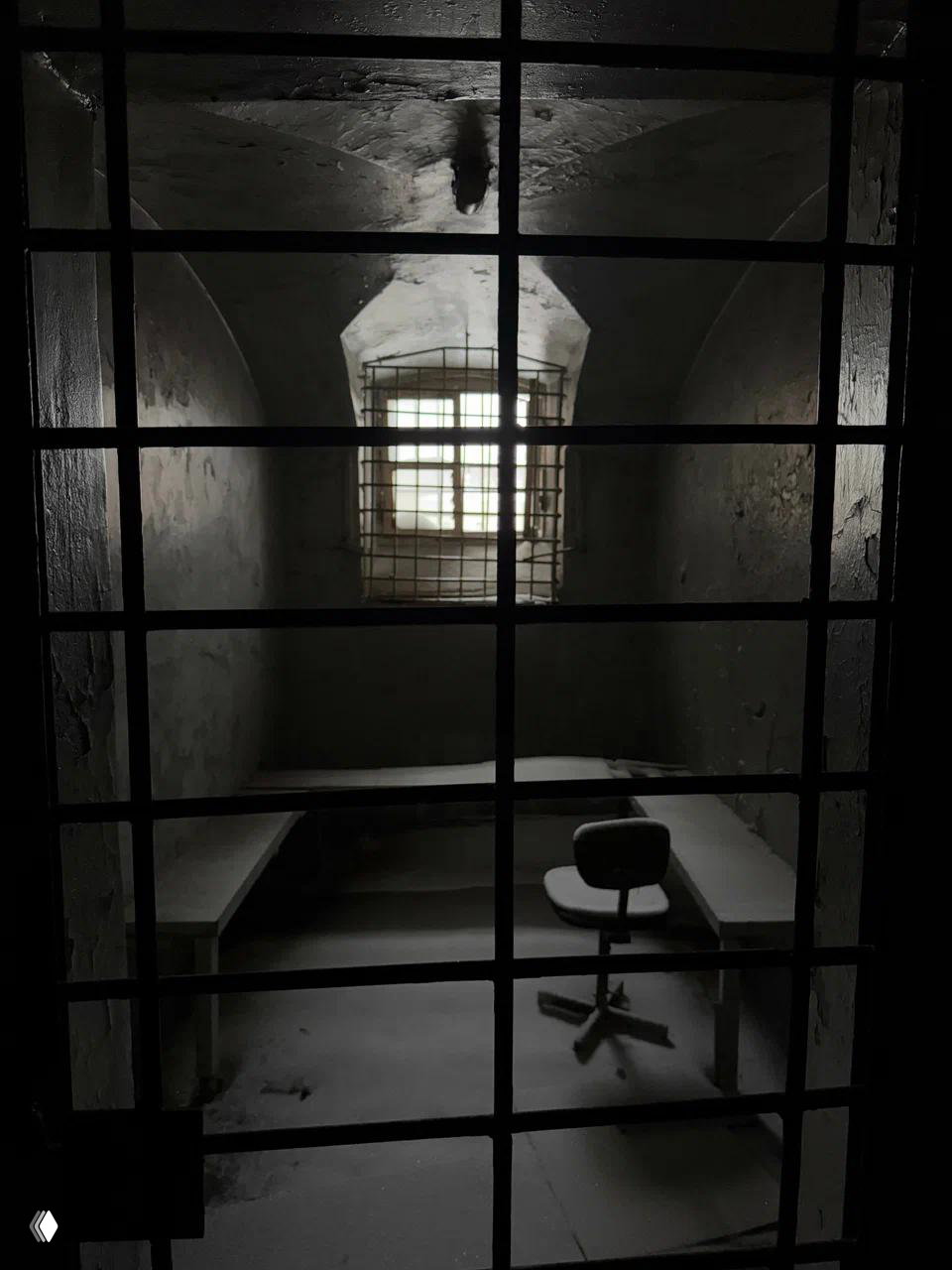 Интерьер одиночной камеры в тюрьме «Кресты»: стол, вращающееся кресло и маленькое решётчатое окно, через которое проходит тусклый свет.