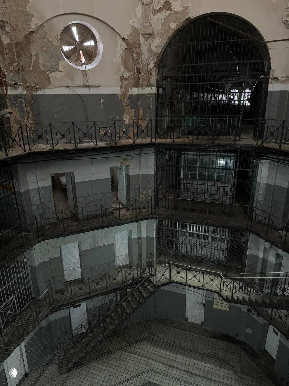 Вид на многоярусный внутренний двор «Кресты»: пустые дверцы камер, металлические балконы, сетки и потертая плитка на полу центрального корпуса.