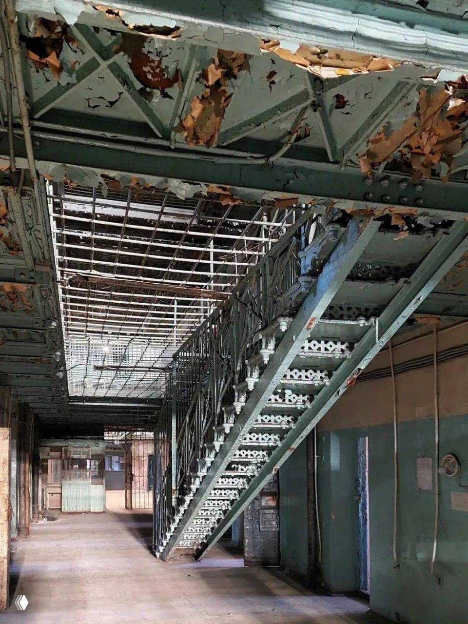 Лестничный пролёт и коридор внутри тюремного блока «Кресты»: облупившаяся краска, чугунные ступени и металлические ограждения под старой кровлей.