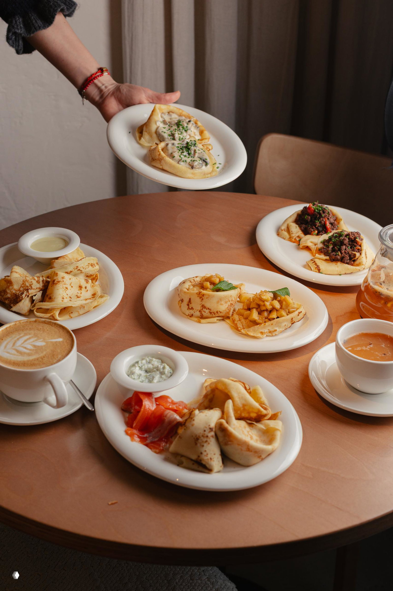 Тарелки с блинами разной подачи, чашка кофе и соусники на деревянном столе; человек подаёт блюдо — масленичная сервировка в бистро.