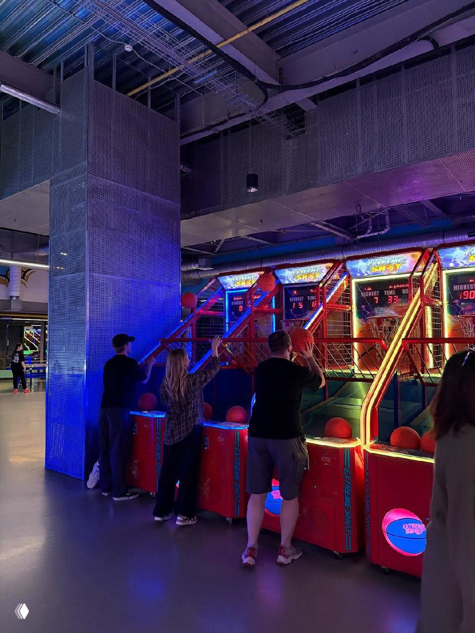 Игровая зона с автоматами для бросков мяча и яркой разноцветной подсветкой: посетители играют в аркадные баскетбольные автоматы внутри комплекса.