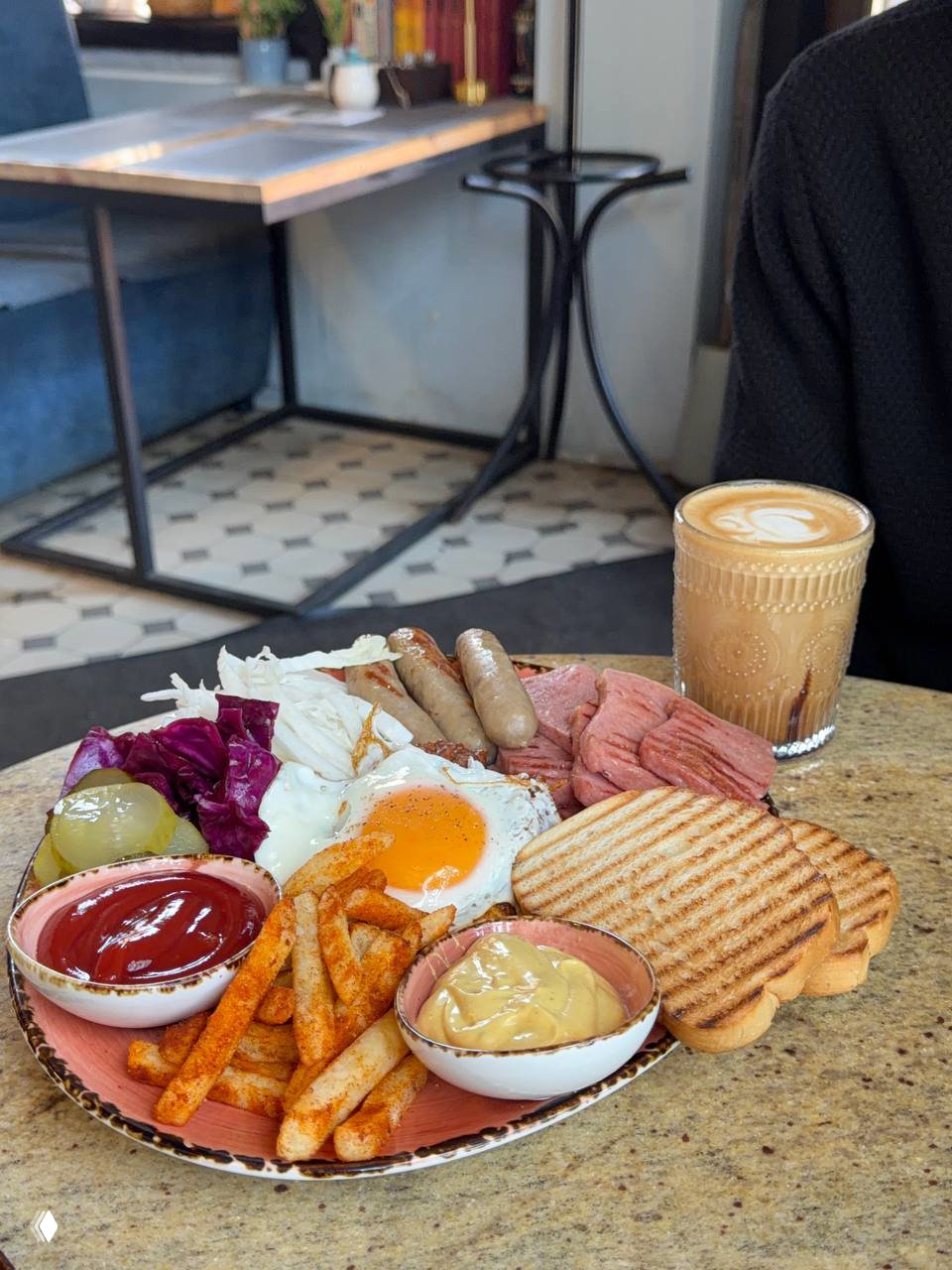 Тарелка с баварским завтраком: яйцо, сосиски, мясные ломтики, тосты, картофель фри и соусы; рядом стакан авторского кофе на столе в кафе.