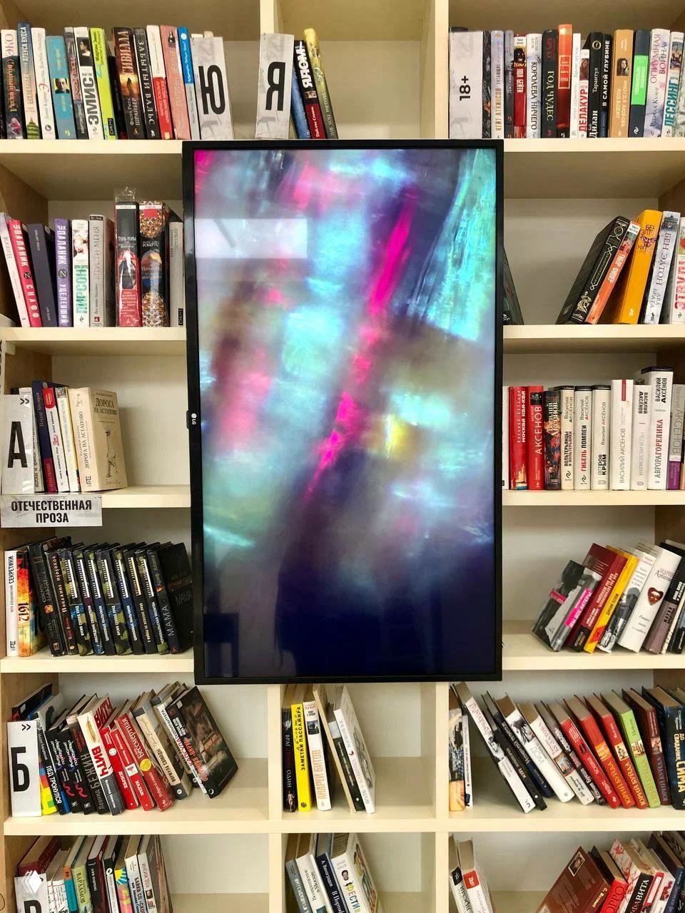 Книжные полки вокруг крупного вертикального экрана с абстрактной цветной графикой — сочетание медиа-инсталляции и фонда книг в центре.