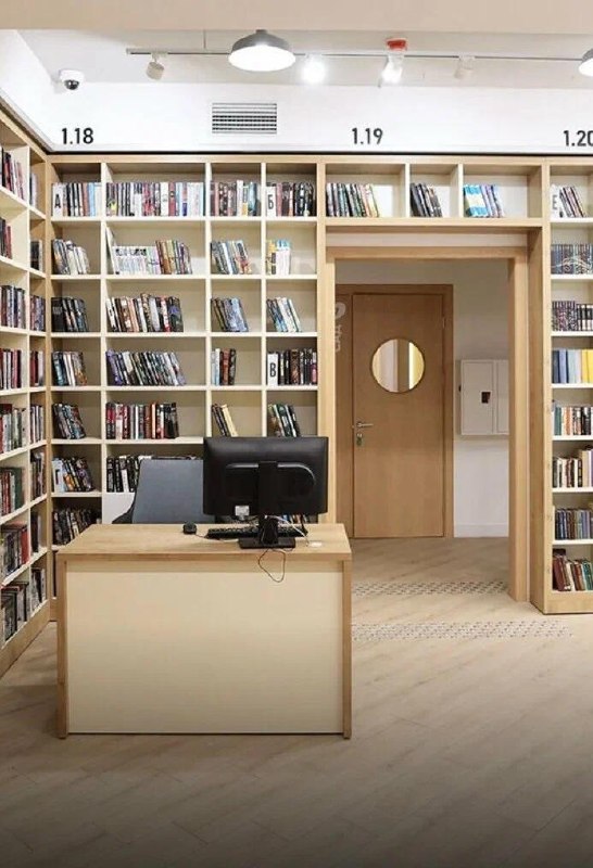 Ресепшен или рабочая стойка на фоне высоких книжных стеллажей с маркировкой секций; светлый интерьер и дверной проём в зал.