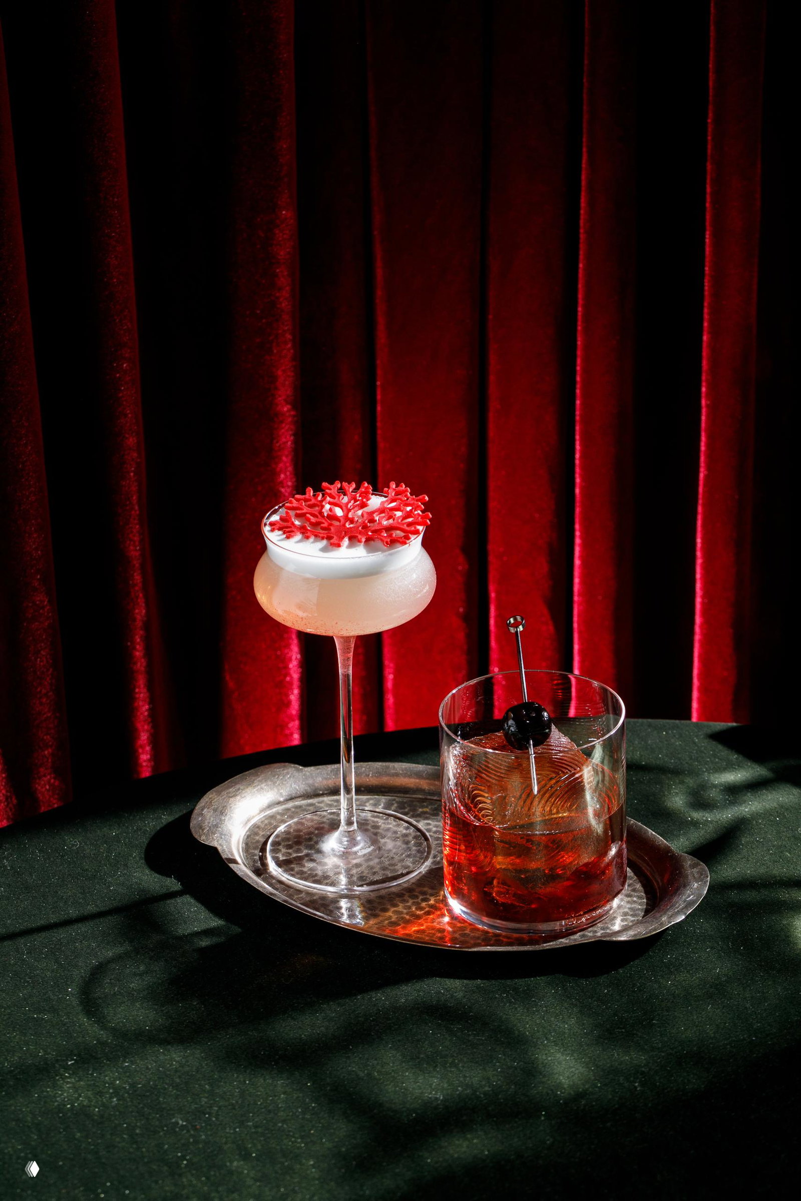 Фото двух коктейлей на серебряном подносе: лёгкий коктейль с белой пеной (Dolce) и тёмный Amaro на фоне бордовых бархатных штор.