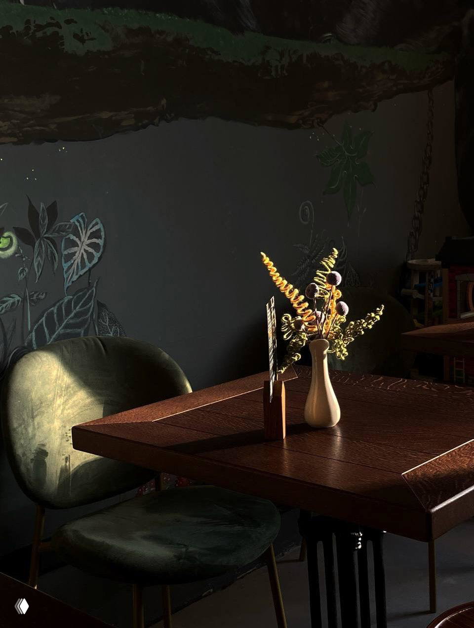 Близкий план деревянного стола с вазой сухоцветов и бархатными зелёными стульями на фоне тёмной стены — деталь уюта и света внутри кофейни.