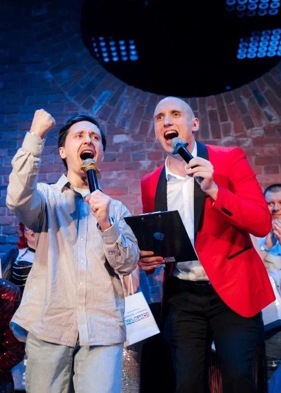 Два ведущих на сцене держат микрофоны и листы с результатами, эмоционально объявляют победителей конкурса перед публикой.