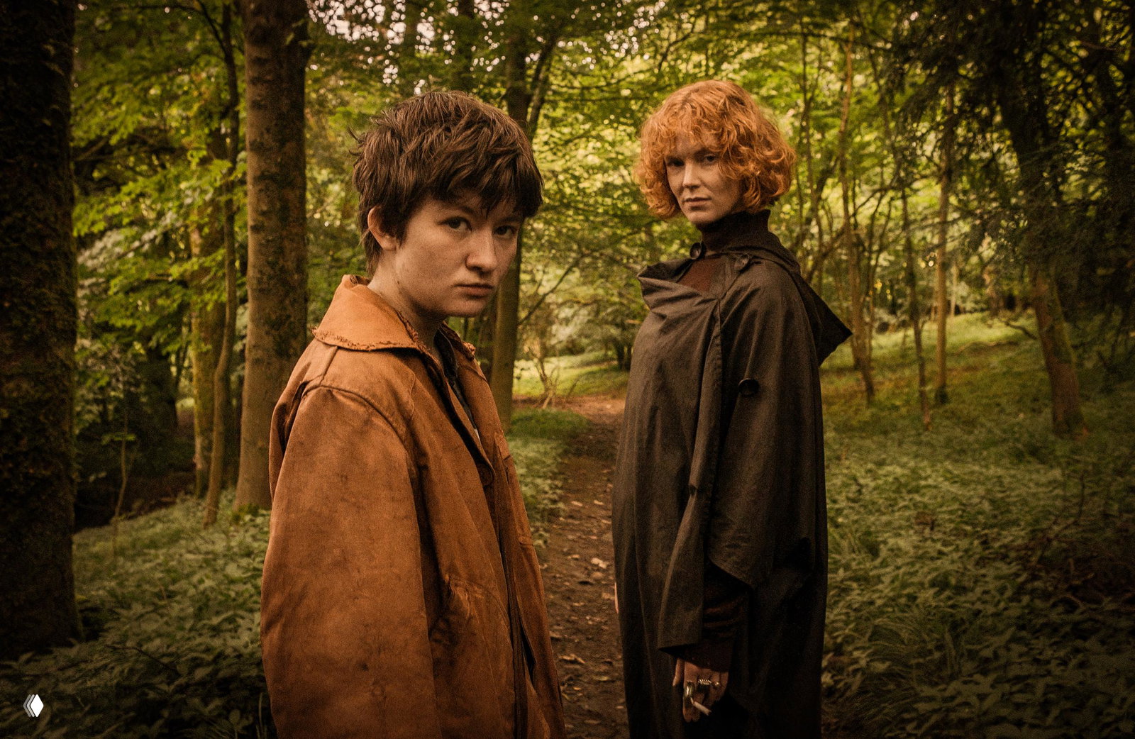 Кадр в лесу: двое персонажей стоят на тропинке среди плотных зелёных деревьев; костюмы и цветокор создают ощущение кельтского фольклора и лёгкой тревоги.