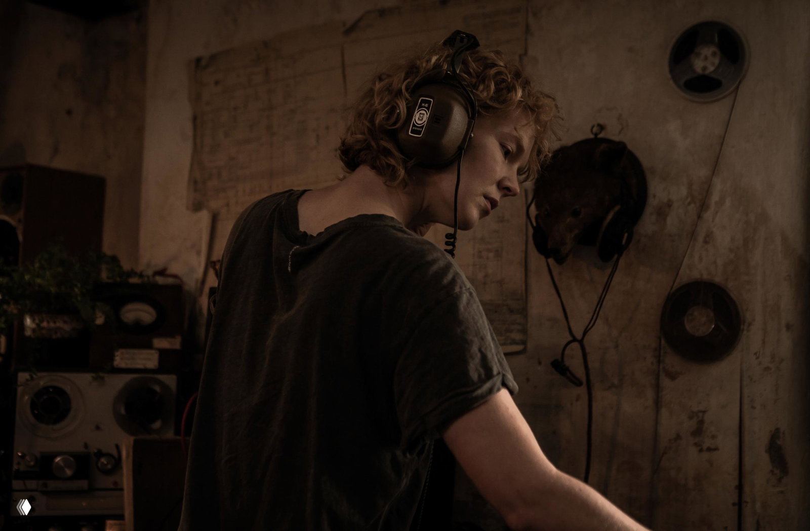 Кадр из фильма: молодая женщина в больших наушниках сосредоточена над ретро-аудиоаппаратурой в плохо освещённой комнате, пыльная техника и атмосферный звук вокруг.