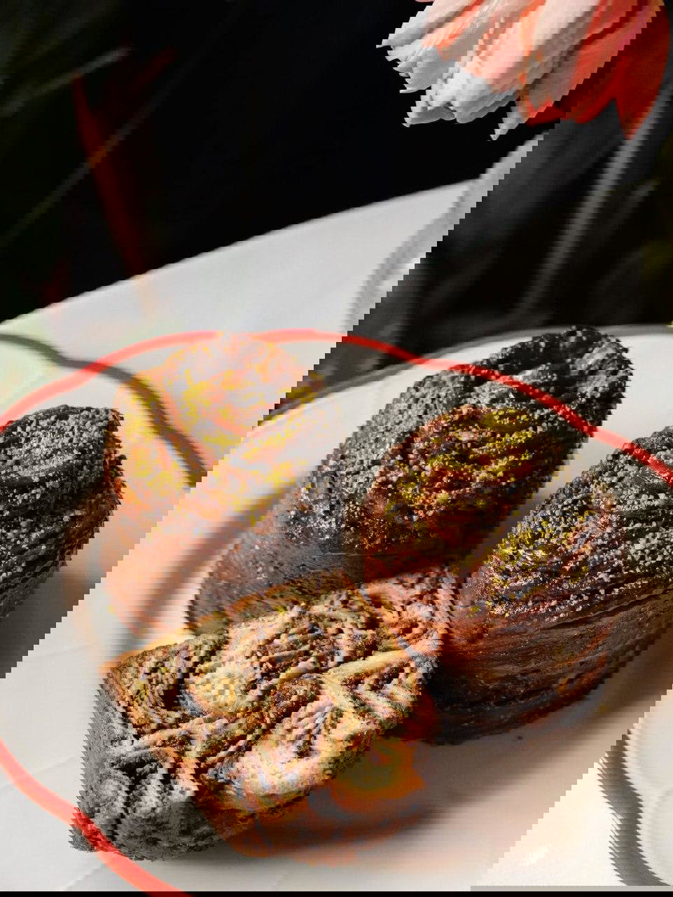 Две шоколадные булочки с посыпкой из фисташек и срезанная часть на тарелке — слоистая структура теста и блеск глазури.