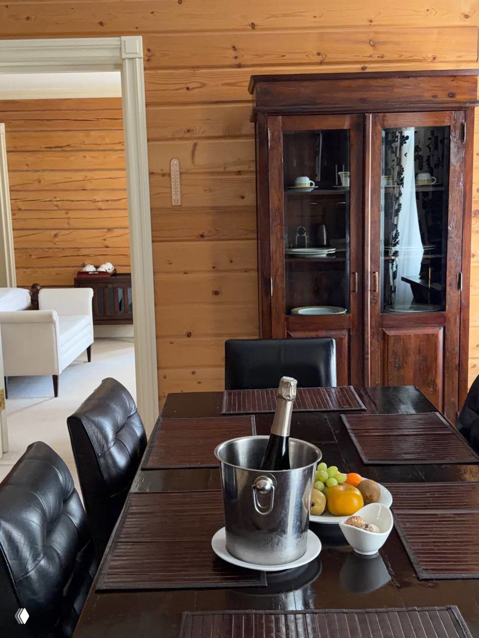 Столовая в деревянном домике: тёмный обеденный стол, ведёрко со шипучим, фрукты и винтажный шкаф на фоне деревянных стен — атмосферный интерьер для завтрака.