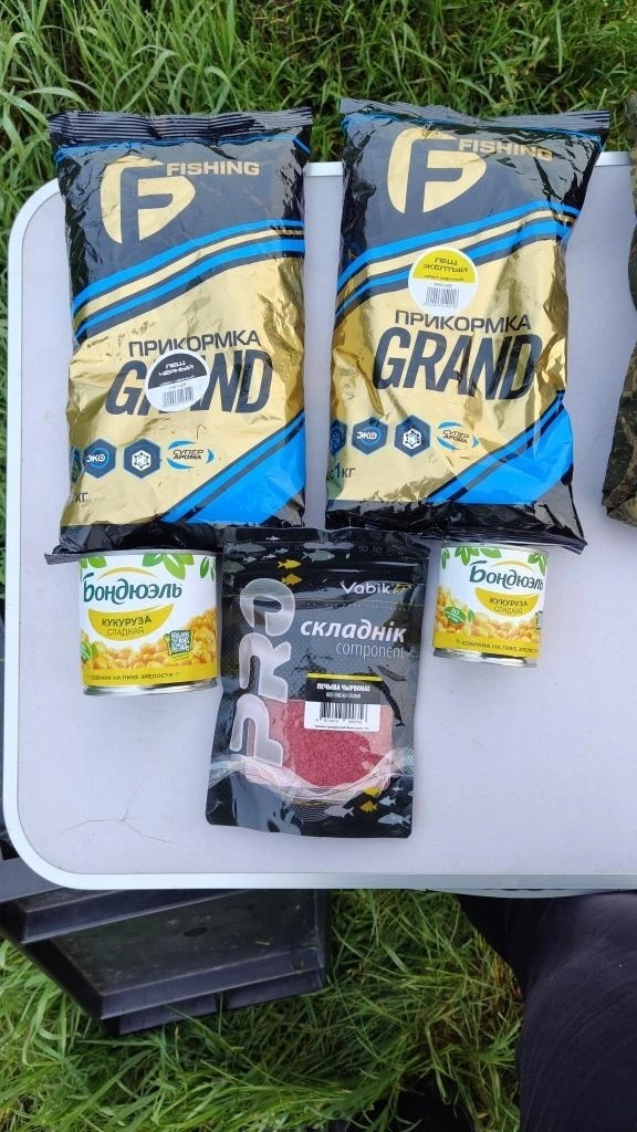 Две пачки прикормки Grand и банки кукурузы и пакет микропеллета, выложенные на столике перед рыбалкой.