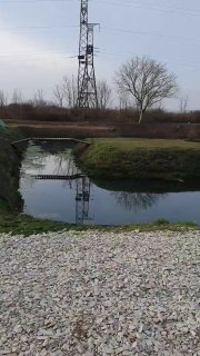 Пруд форелевого комплекса в Абинске: зеркальная вода, отражение опоры ЛЭП, гравийный берег и голые деревья на фоне неба.