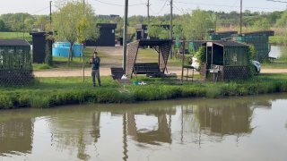 Панорамный вид на берег платного пруда в Адыгее: рыболов у воды, причалы и беседки, прибрежная зелень и отражение построек в спокойной глади воды.