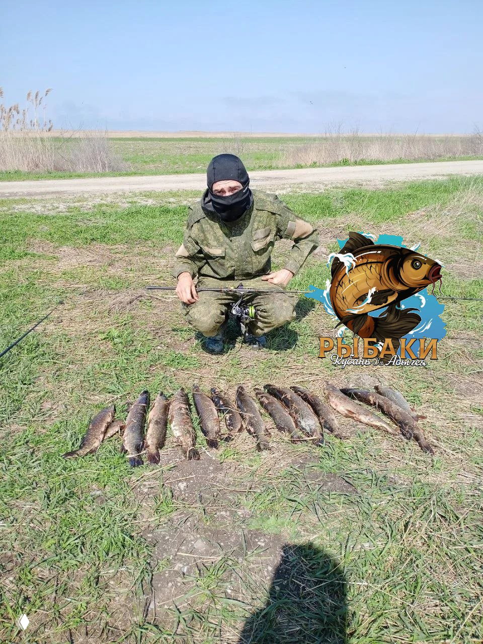 Рыбак в маске и камуфляже сидит рядом с рядом пойманных щук на земле у дороги, видна рыболовная катушка и логотип канала «РЫБАКИ».