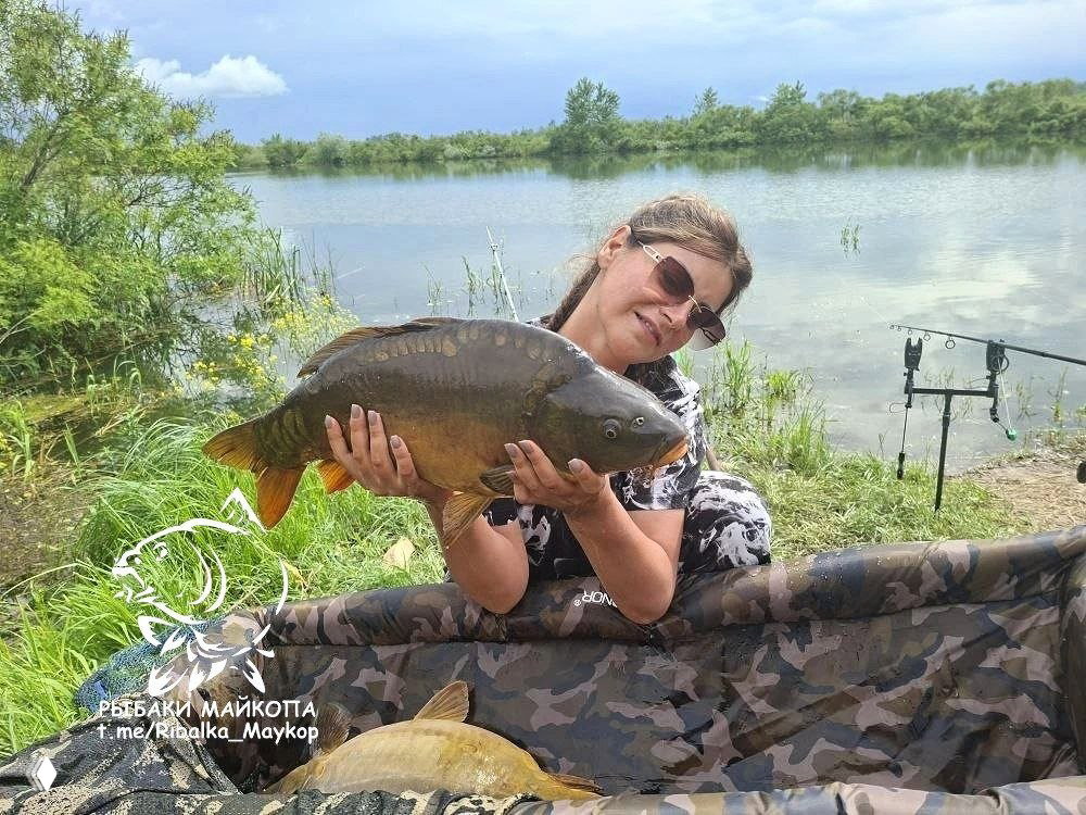 Рыбачка показывает зеркального карпа в мате для взвешивания, на заднем плане рогулька и спокойная поверхность пруда под облачным небом.