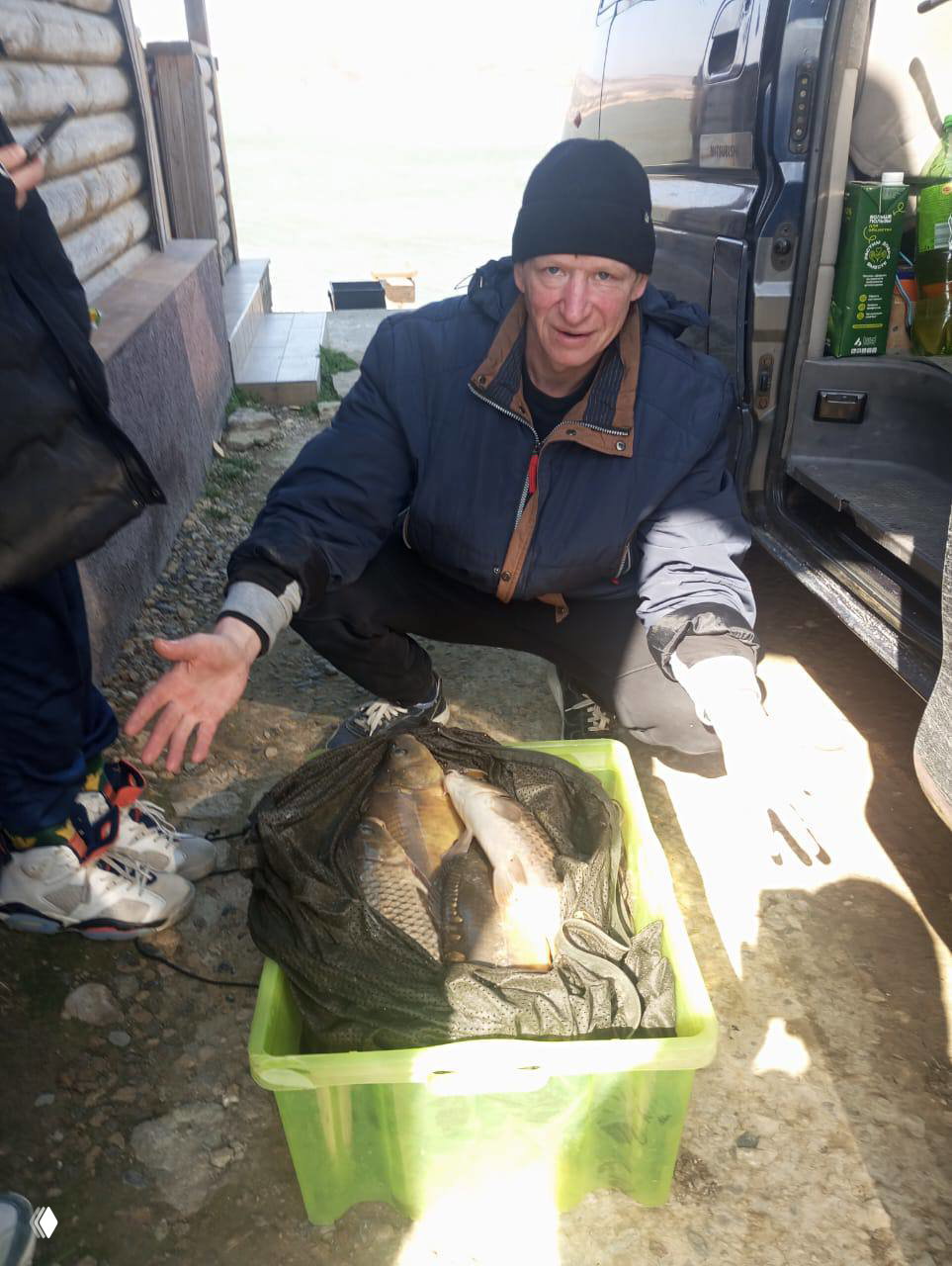 Мужчина в куртке присел рядом с фургоном; в пластиковой коробке лежит несколько карпов — улов с Красной Горки.