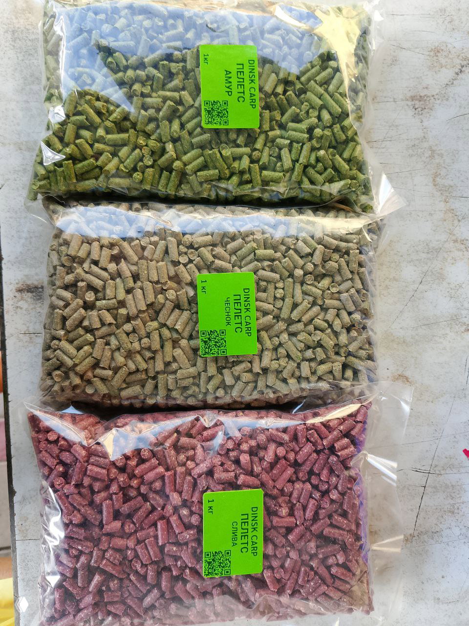 Три прозрачных пакета с пеллетс-прикормкой DINSK CARP разных вкусов: зелёный, бежевый и розоватый, на столе сверху.