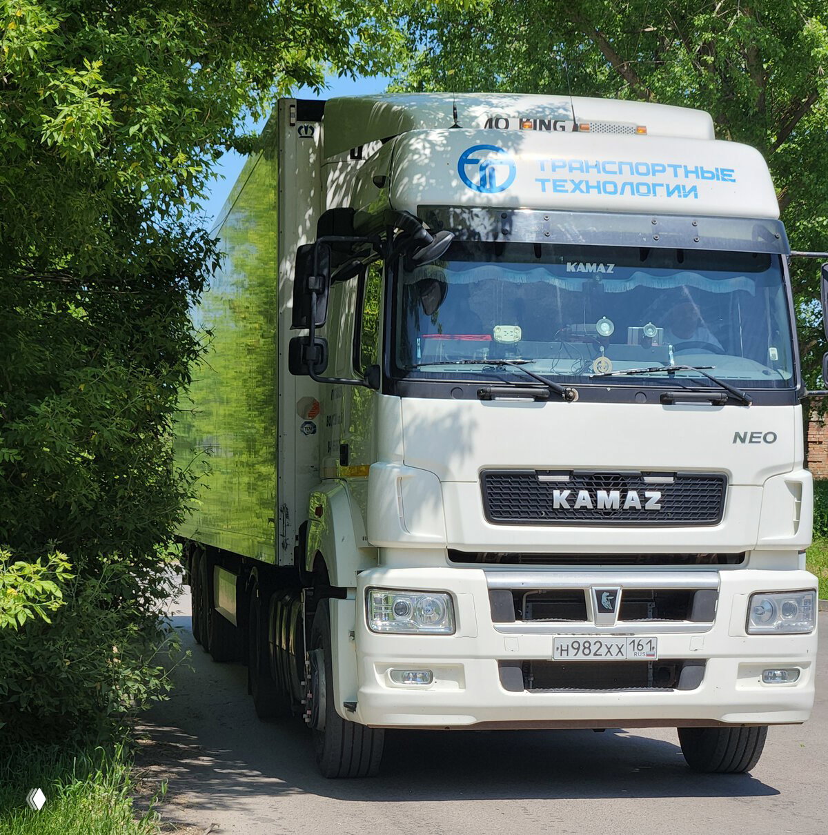 Белый тягач KAMAZ с бортовым прицепом и надписью «Транспортные технологии» на капоте, припаркованный у зелёных деревьев; вид спереди, фокус на кабинe.