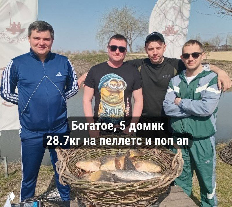 Групповое днёвое фото: четыре рыбака позируют за корзиной с несколькими карпами на берегу пруда, видна подпись о 28.7 кг и флаги пруда.