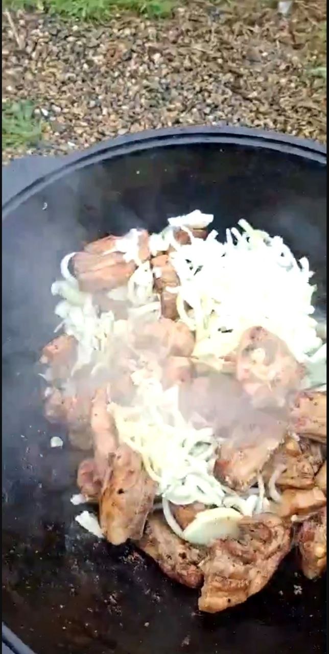 Казан с жарящимся мясом и луком на открытом огне у пруда — кадр ужина для участников соревнований, виден пар и процесс приготовления еды.