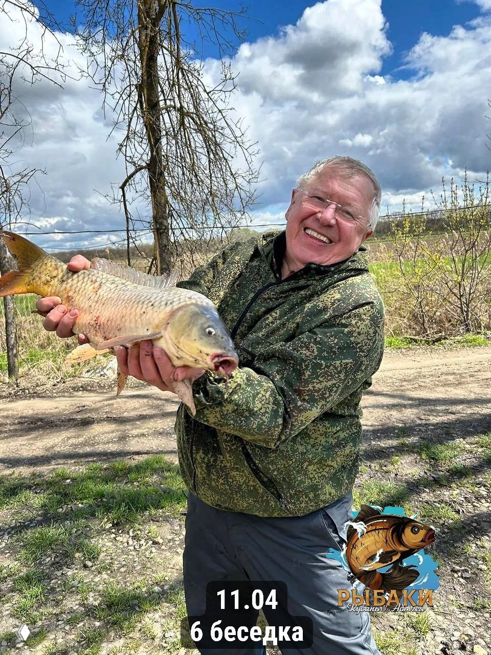 Пожилой мужчина улыбается, держа большую рыбу на открытом участке у дороги; на фоне деревья и облачное небо, видна надпись с датой.